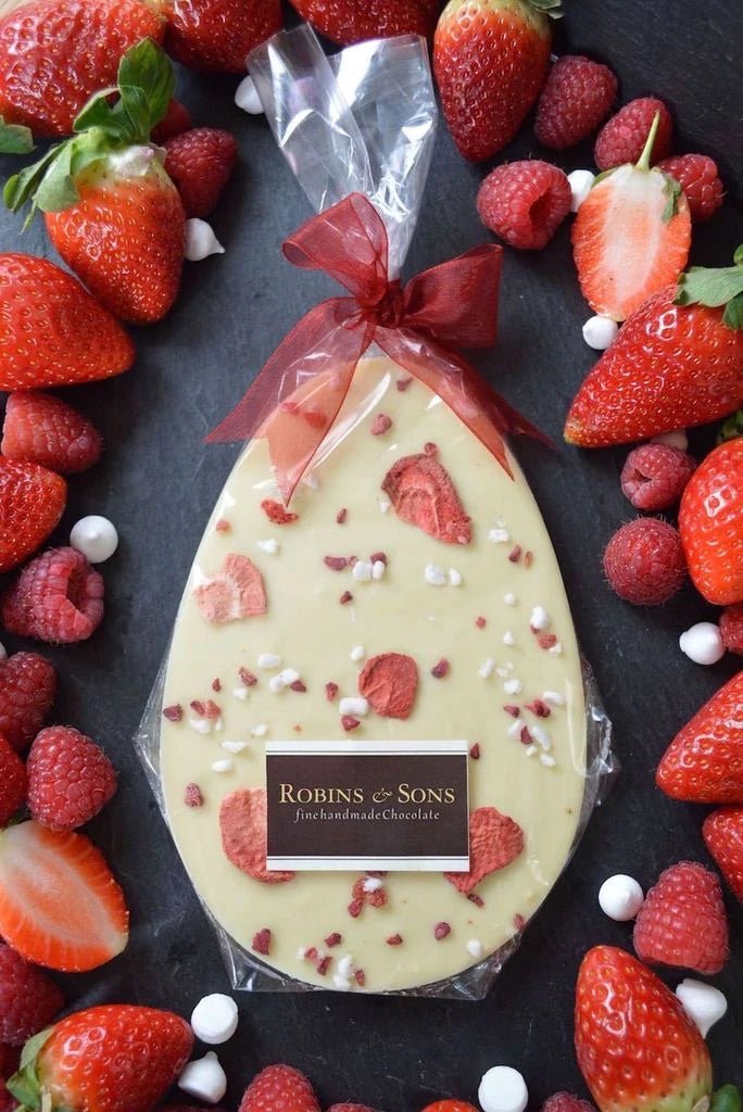 Very Berry Easter Egg. White oval-shaped chocolate bar topped with strawberries and meringue, in clear packaging and tied with a red bow.